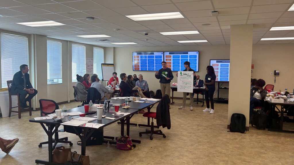 Students and adults in a large room with long tables and a whiteboard; some people are seated while others are standing.