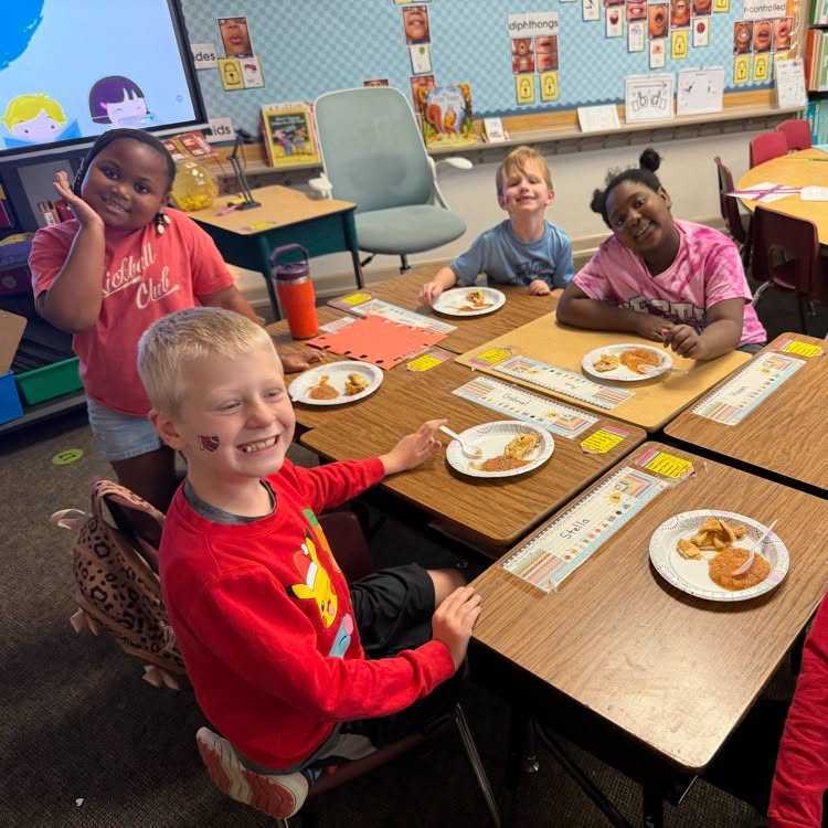 Children seated around a table, each with a plate of food, smiling and talking, in a classroom.