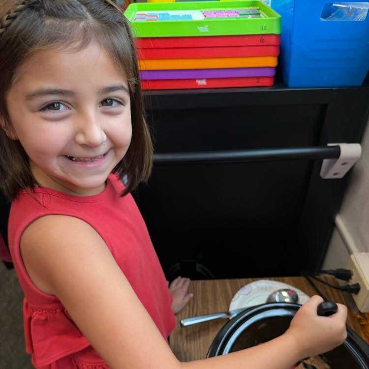 Girl in red shirt smiling, holding a pot, in a classroom with storage boxes behind her.