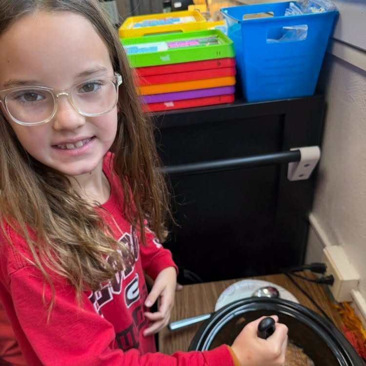 Girl with glasses holds a slow cooker in a classroom with a shelf of colorful bins behind her.