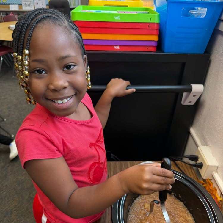 A smiling child holding a ladle stirs a slow cooker full of food. Behind her are a black filing cabinet and a shelf with colorful folders.