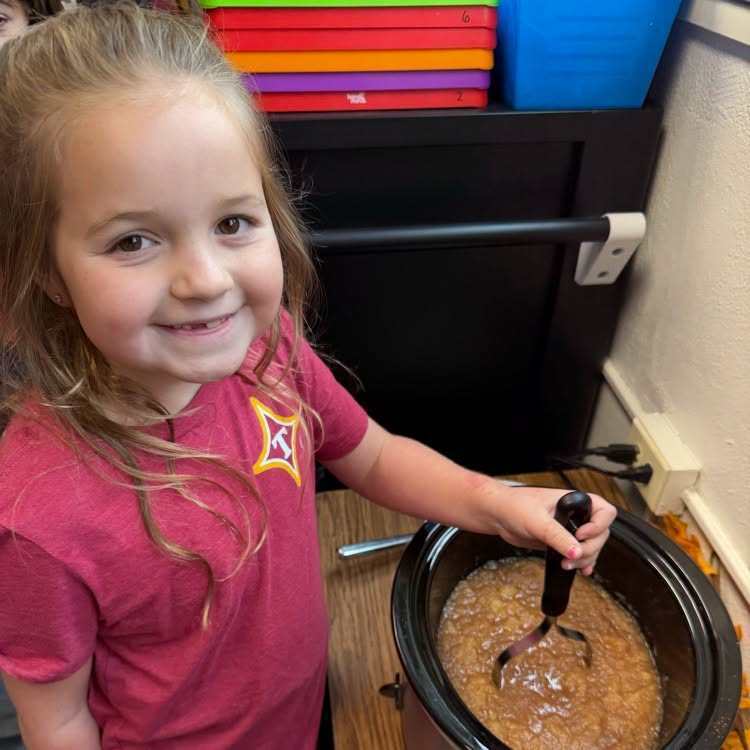 A young girl in a red shirt smiles as she stirs a pot of food with a ladle.