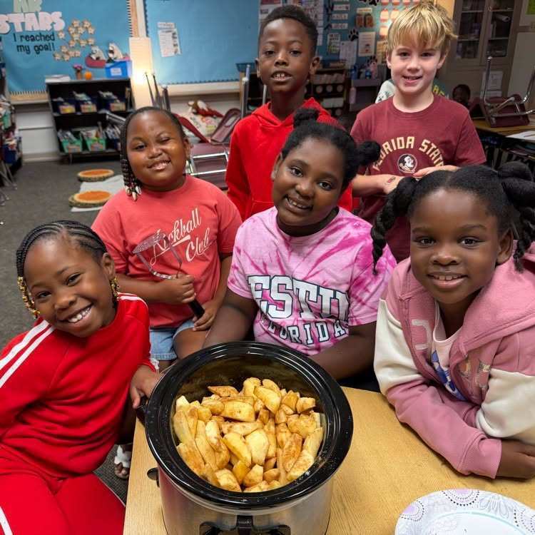 Group of children posing next to a crockpot of food on a table in a classroom.