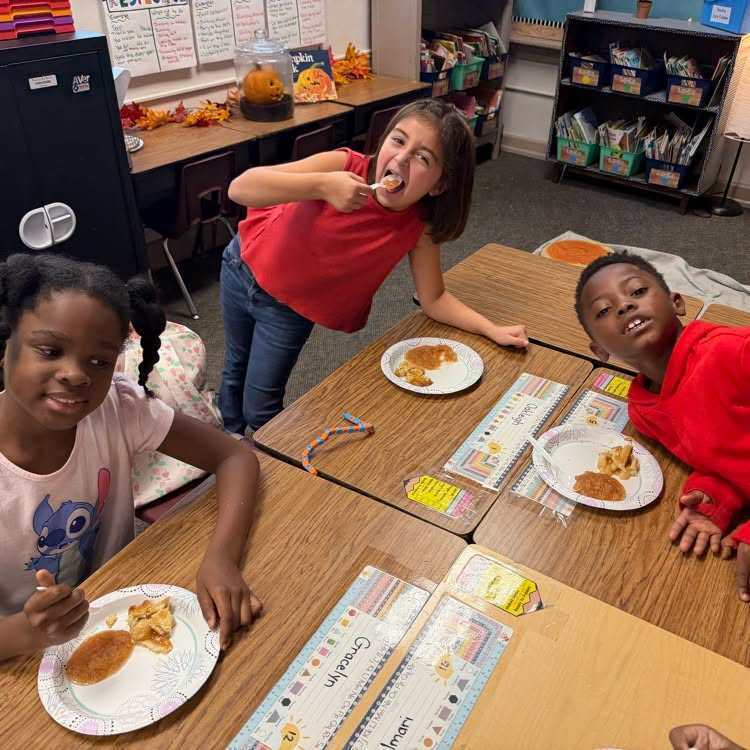 Three students eating pancakes and smiling at a desk; a shelf with books is in the background.