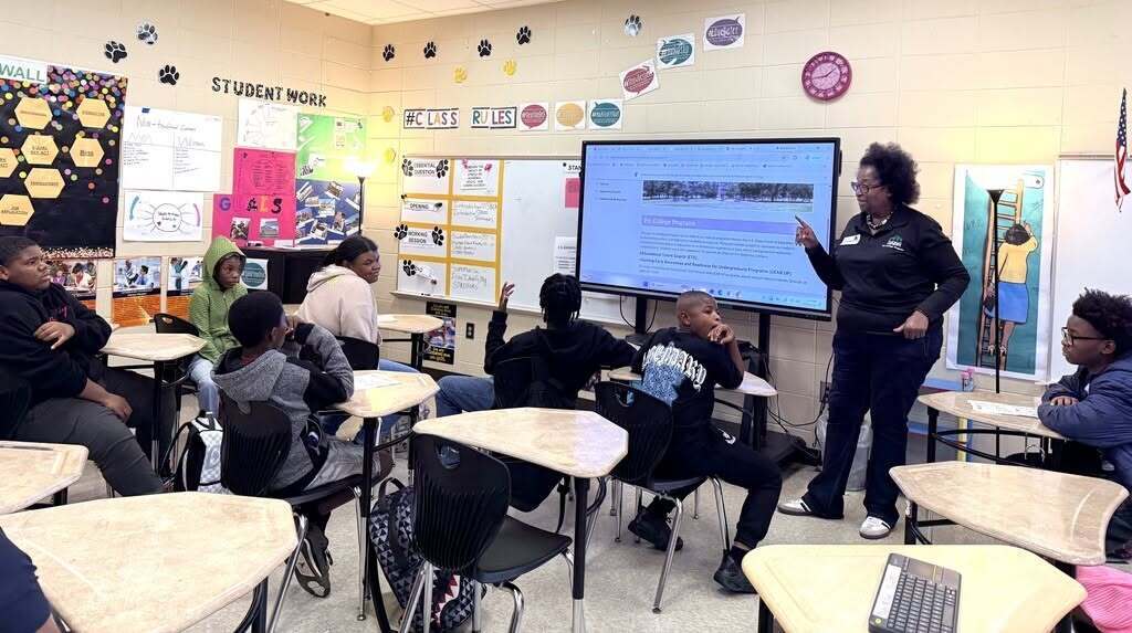 A teacher stands in front of a projector screen in a classroom with students seated at desks.