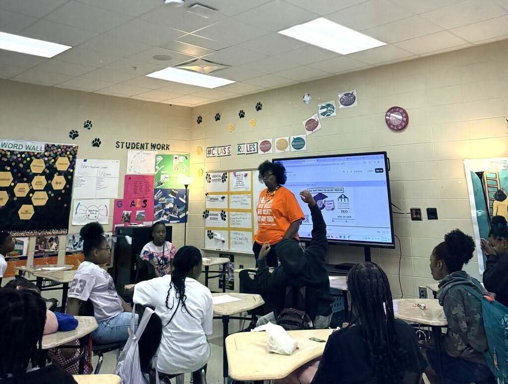 People seated at tables in classroom. Person in orange shirt stands at front with projector screen behind them.