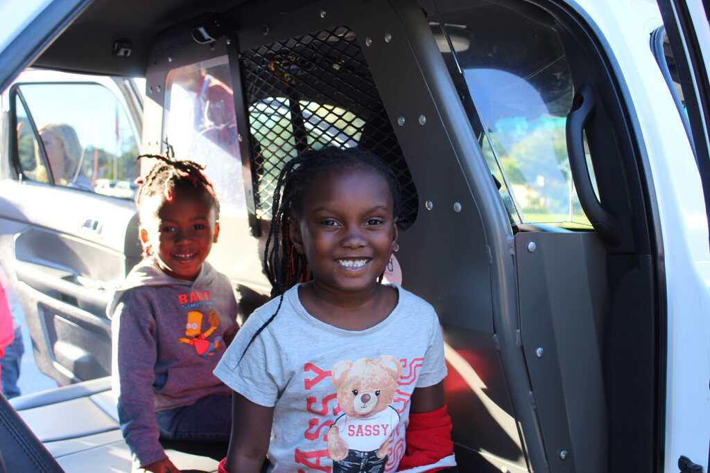 Two children sit inside a vehicle; one smiles. One girl wears a purple shirt; the other wears a white shirt with a bear.