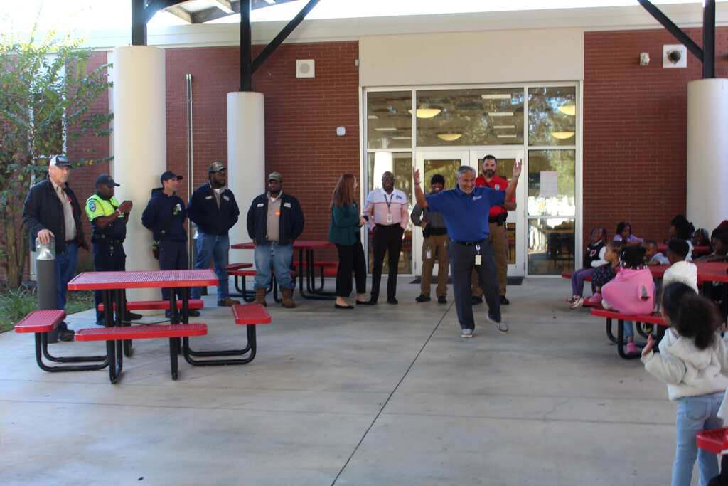 Adults standing outside school near tables, with some seated on benches, with children seated and standing nearby.