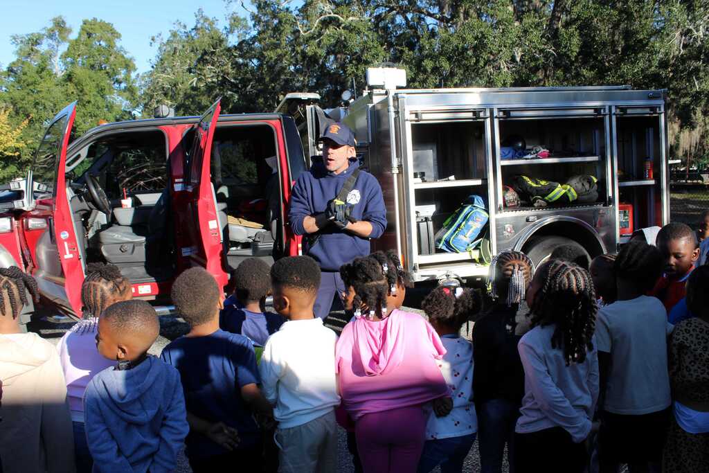 A man explains something to a group of children in front of a red fire truck.