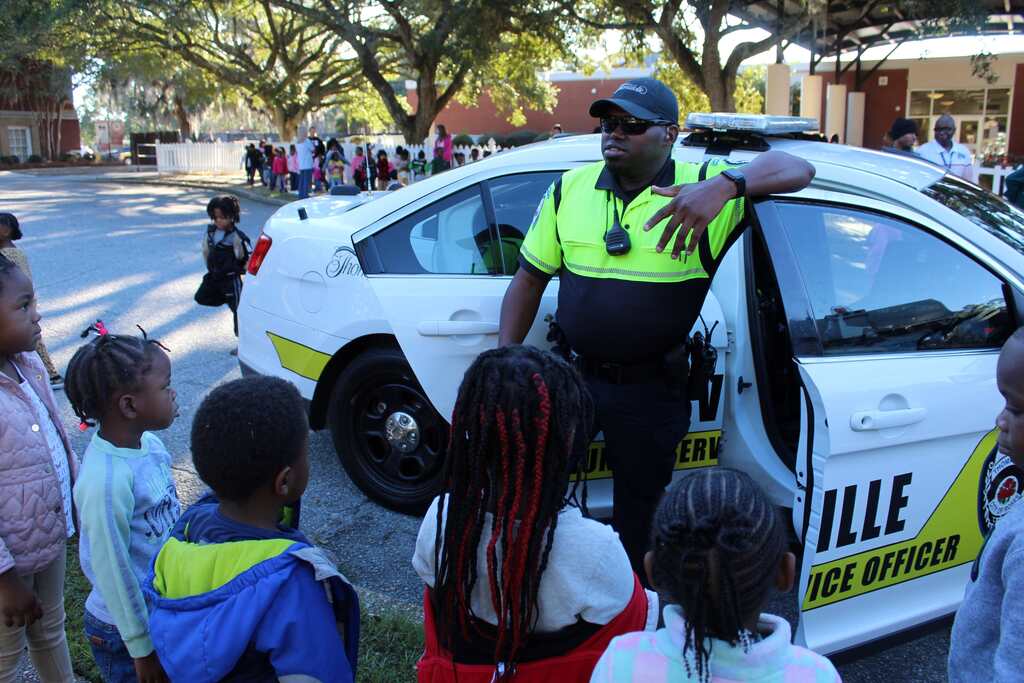 Police officer with hat and watch speaks to children, one with braids, near a police car.
