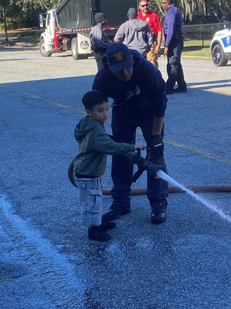 Child holds hose with firefighter; water sprays in parking lot. Vehicles and people in background.
