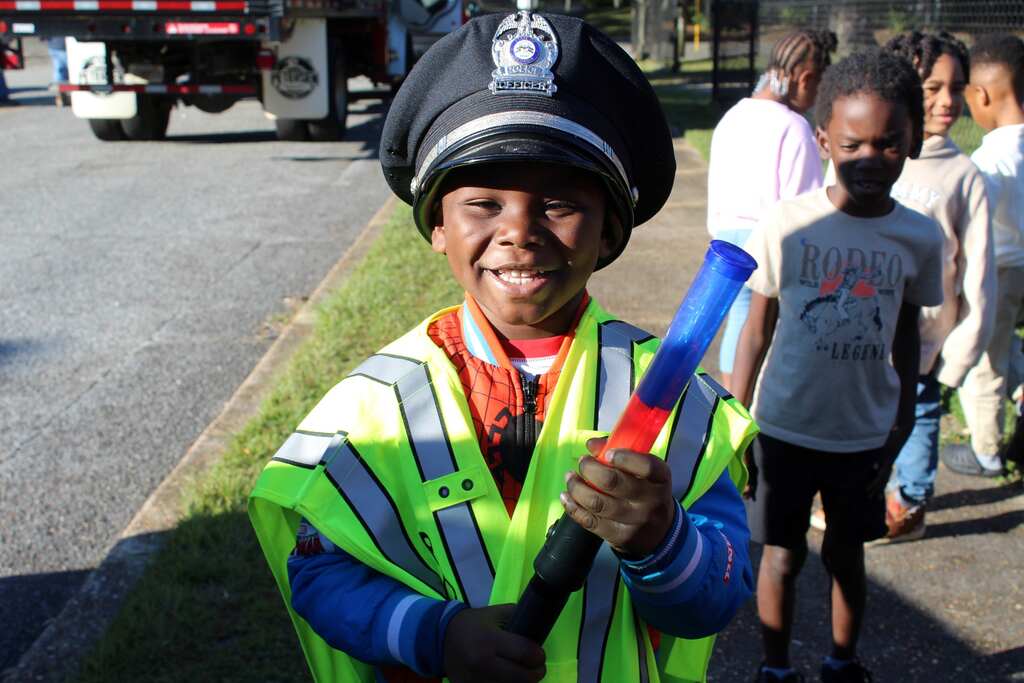 A child in a reflective vest holds a neon-colored baton and smiles. Other children walk behind them.