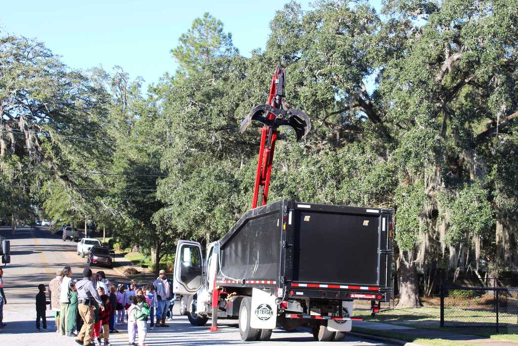 A large truck with a crane on top stands near a road, with several people observing from a distance. Trees and greenery are in the background.