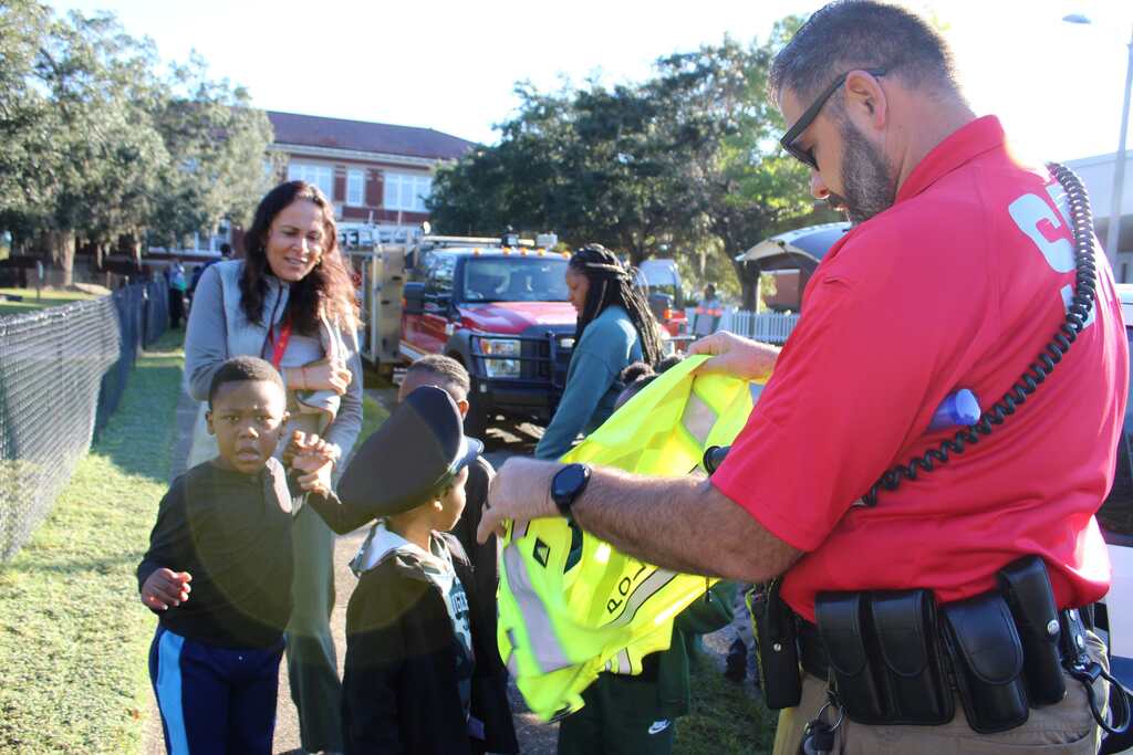Man wearing a red shirt hands yellow vest to two children, one of whom wears a black hat.
