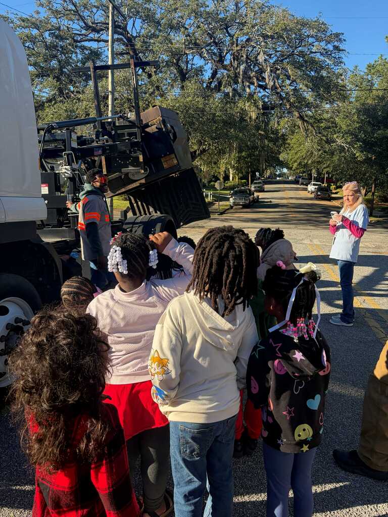 Kids listening to adult in front of large vehicle. Trees, cars, and sidewalk background.
