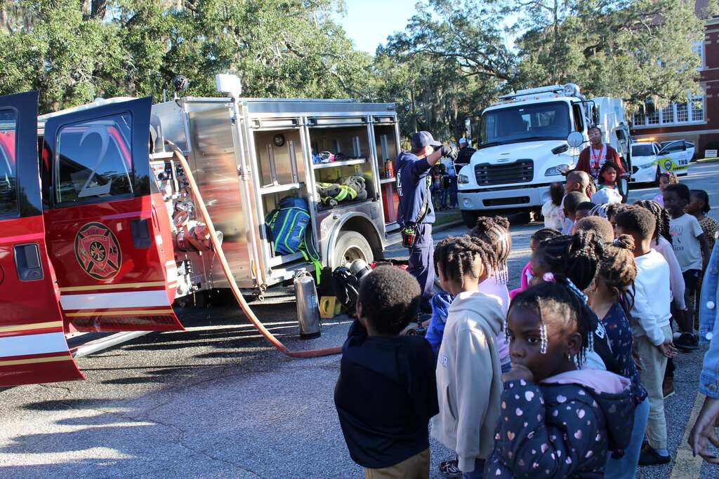 Students stand around a fire truck parked on the street with trees and another vehicle behind.