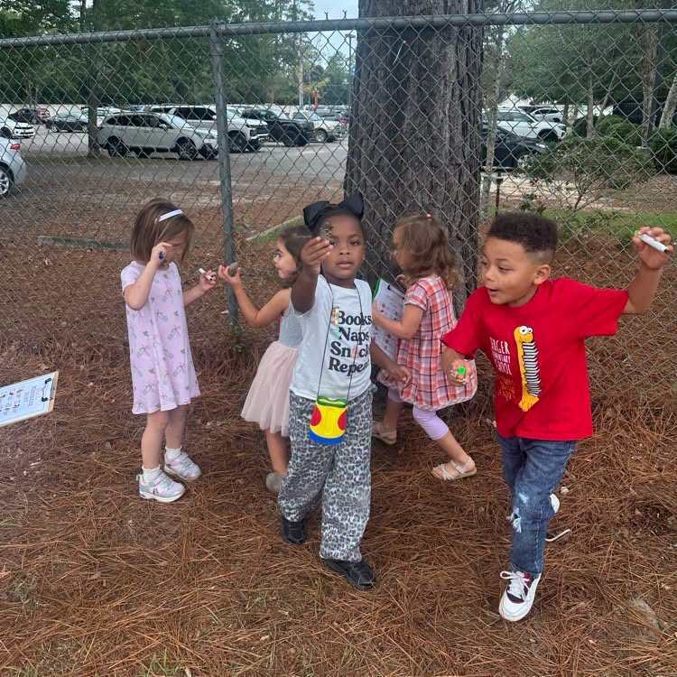 Children outside by a tree, some wearing sneakers, one holding a candy bar, and a fence in background.
