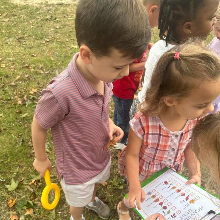Group of children outside. A boy holds a magnifying glass. A girl looks at a paper.