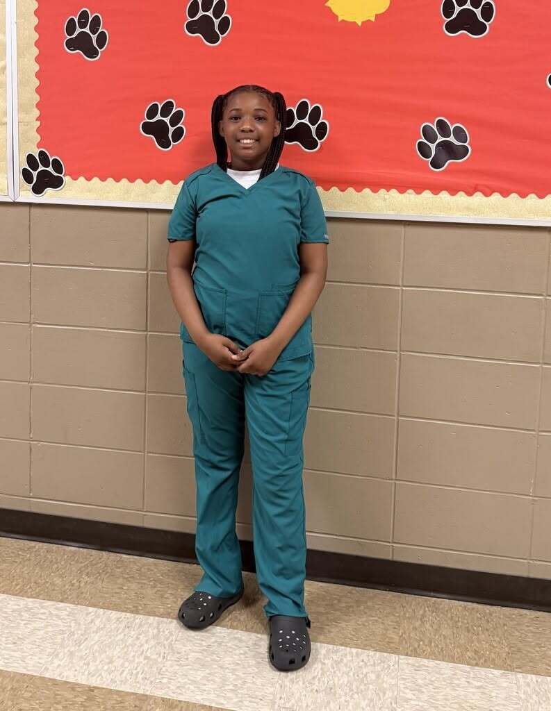 A girl in teal scrubs stands against a wall with paw print decorations. She looks at the camera.