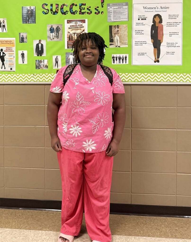 Person in pink scrubs with flowers standing in front of green bulletin board with photos and text.