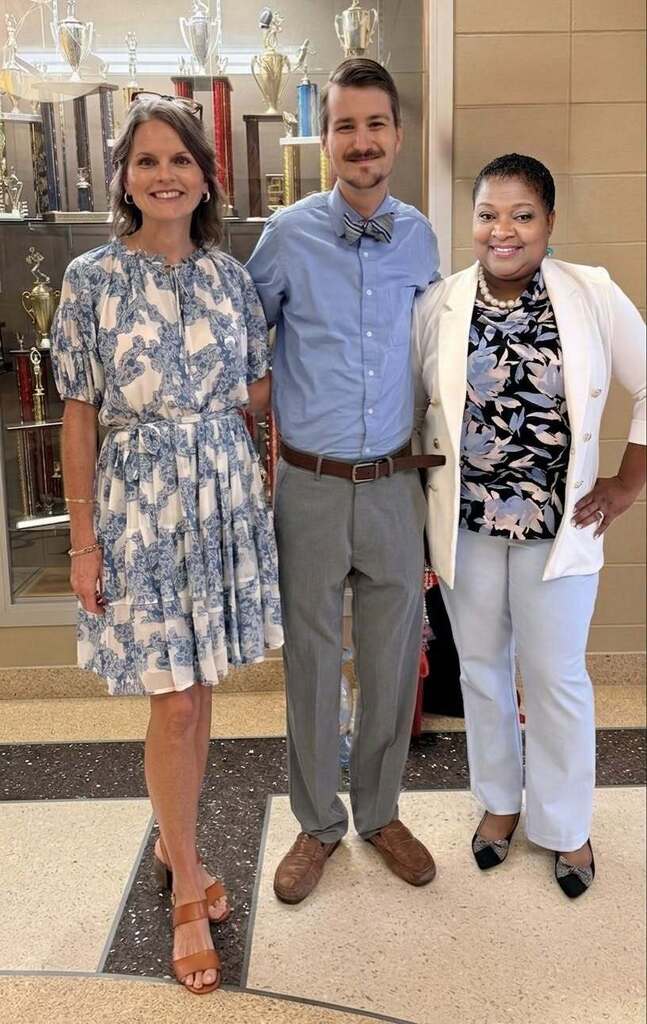Three adults stand together, smiling, in a hallway with tiled floor and display cases with trophies.