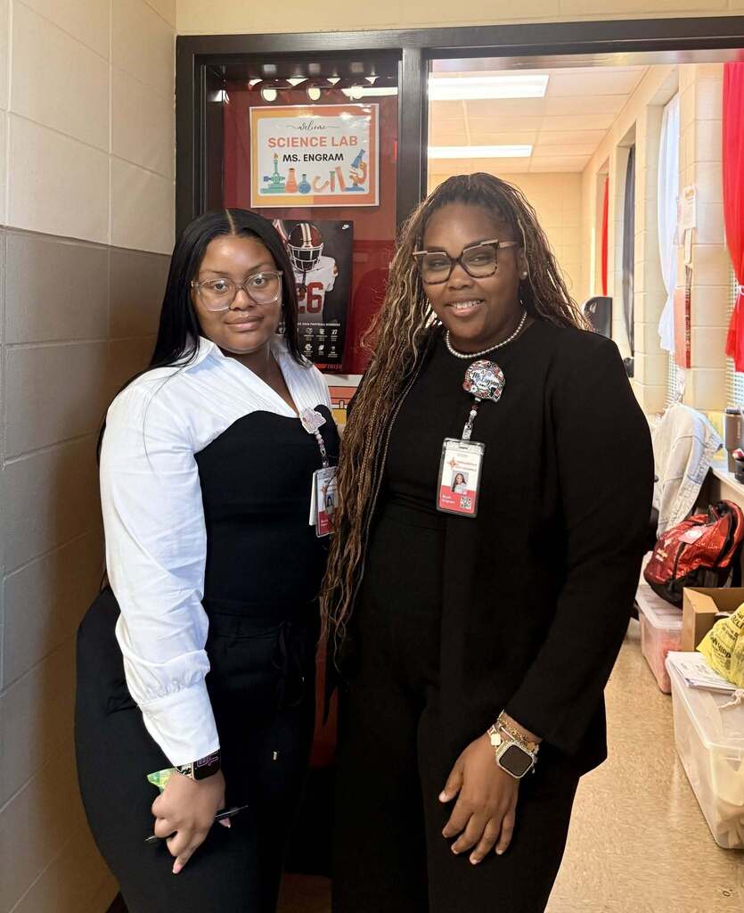 Two women standing next to each other in a hallway, one wearing a white shirt, the other black.
