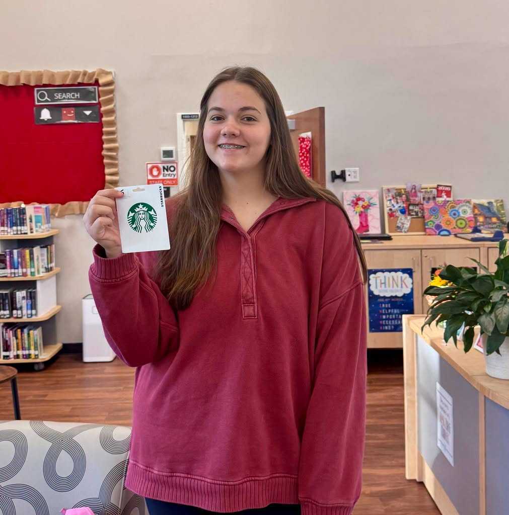 A young girl in a maroon hoodie holds a Starbucks card in front of a library.