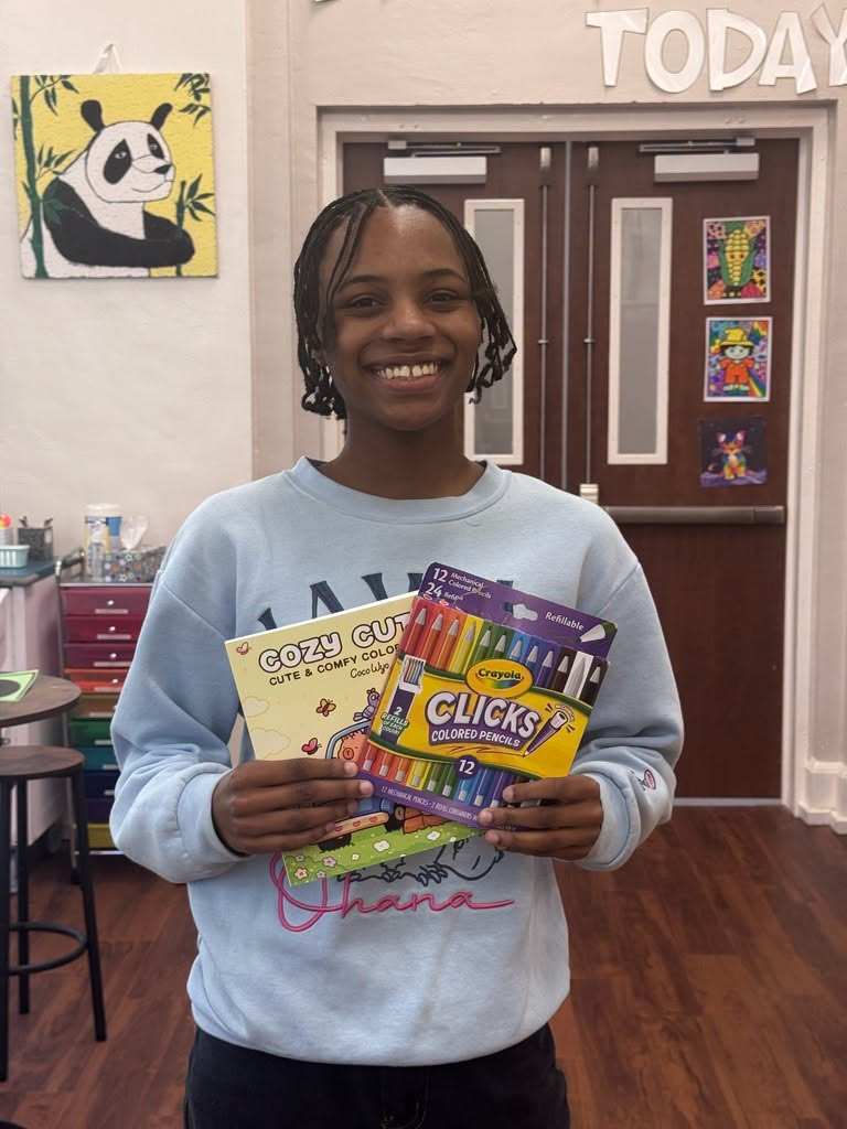 A young person holding coloring books and crayons stands in a classroom with wooden floors and doors.