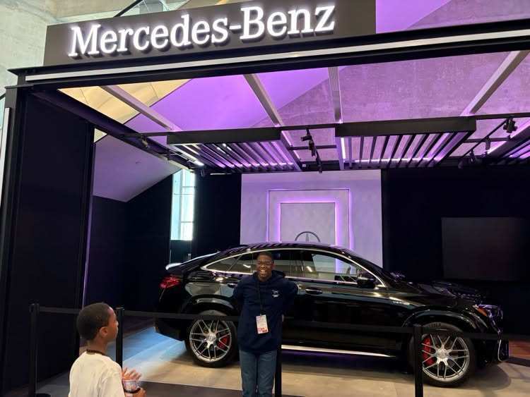 Two people examine a black car inside a showroom with a "Mercedes-Benz" sign above.
