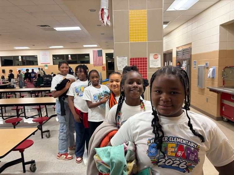 Five girls stand in a school hallway; four wear matching white shirts. The girl in the foreground wears a backpack.