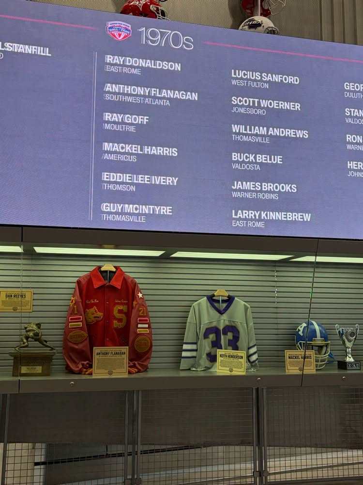 Display of sports jerseys and trophies on a shelf with a large screen showing names and numbers.