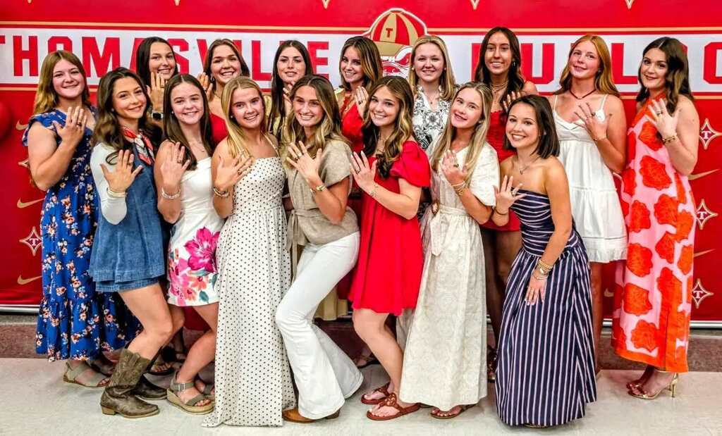 A group of girls poses for a photo in front of a red banner. They wear various dresses and make peace signs.
