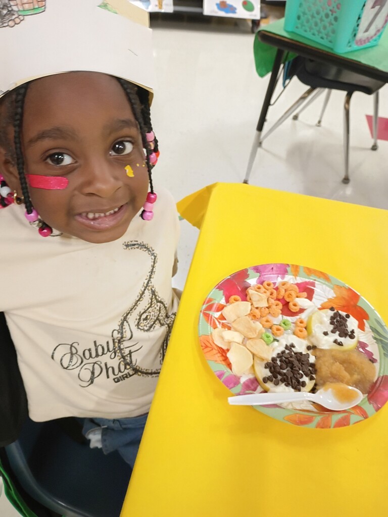 Child sitting at a table with a plate of food, smiling with paint on face.