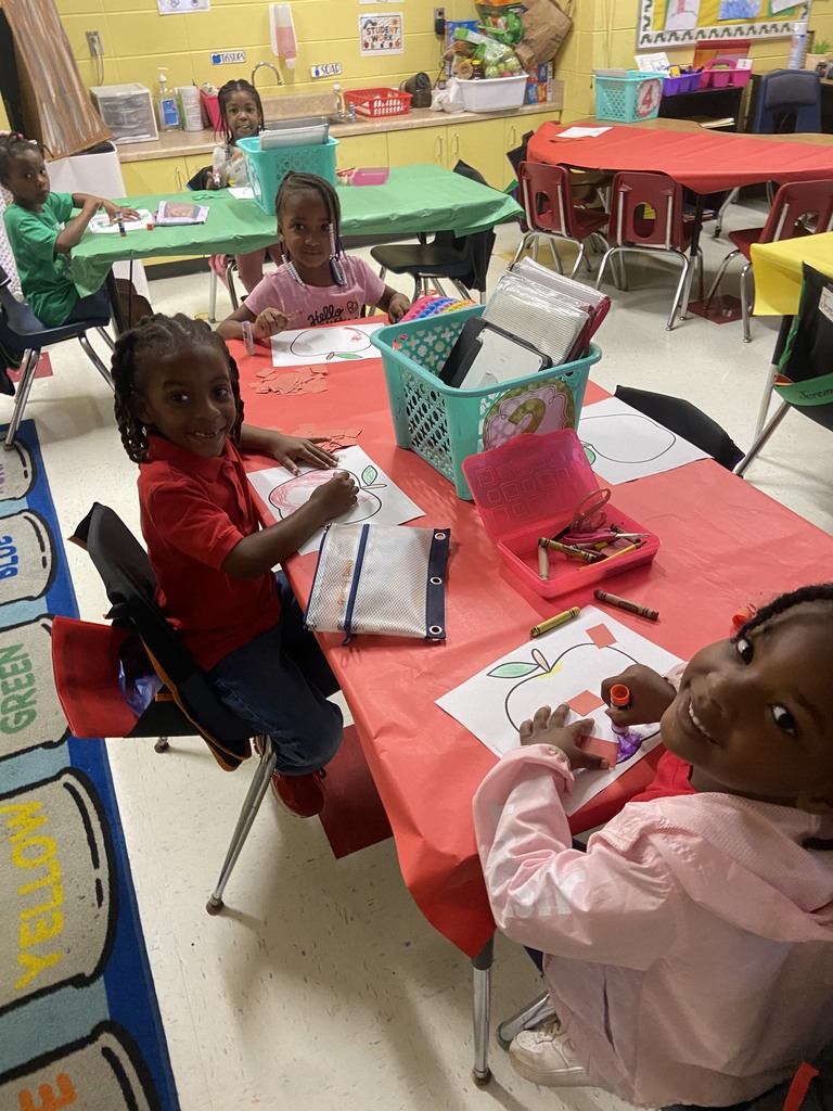 Four children seated at a red table, working on crafts. A girl in a red shirt draws on paper.