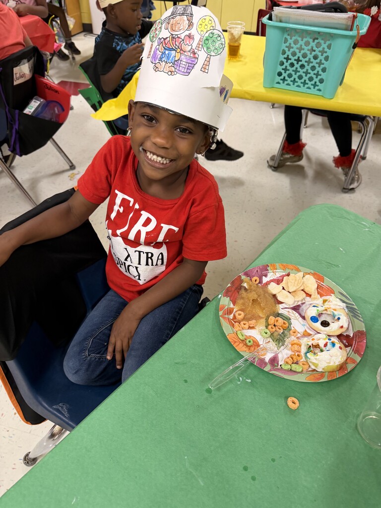 Child wearing chef hat sitting at table with plate of food, other children in background.