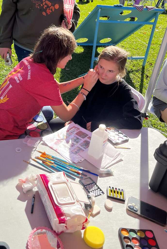 Woman seated at table with supplies for face painting, another woman helps her with makeup.