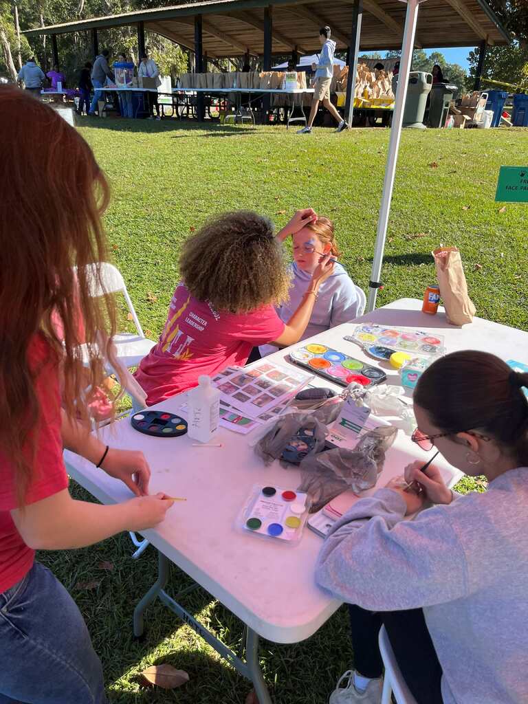 Children and adults paint faces at a table outdoors. A tent and tables with items in the background.