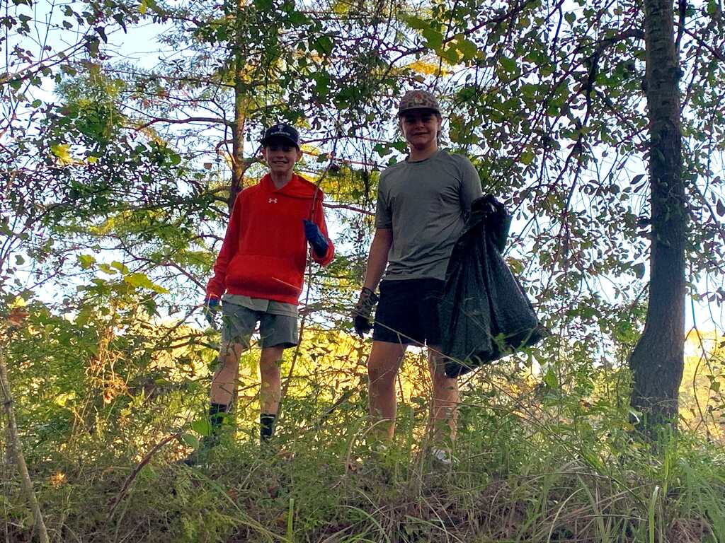 Two people stand in a field, wearing hats, gloves, and carrying trash bags. One wears a red jacket. Trees and greenery are behind them.