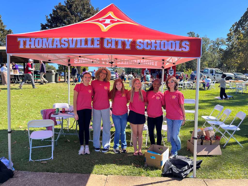 Six people in pink shirts and white sneakers pose under a tent on a grassy field.