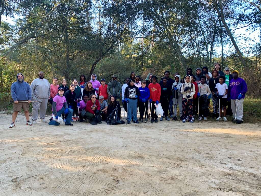 Group of students and staff outdoors, standing in a line, with some holding bags, on a dirt path.