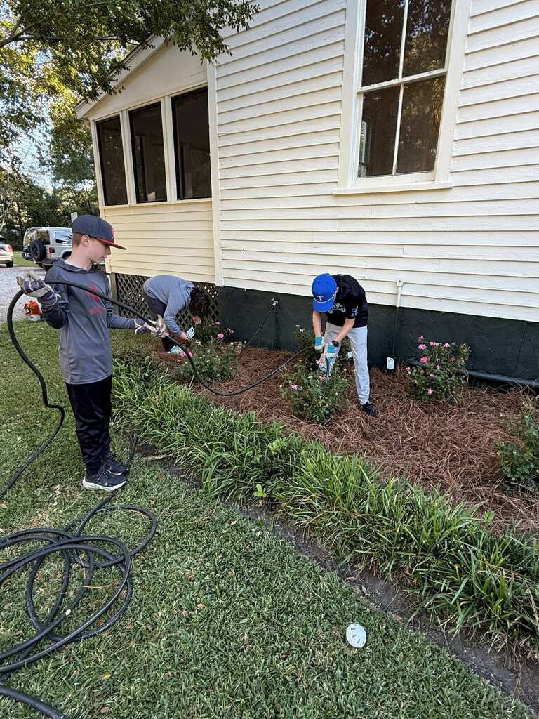 Three students in front of a white house, one spraying water, one planting, one using tools.