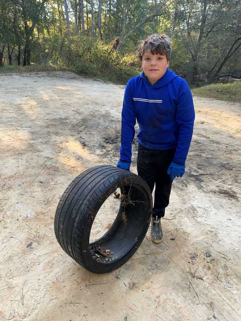A young boy in a blue jacket holds a large tire on a dirt road with trees in the background.