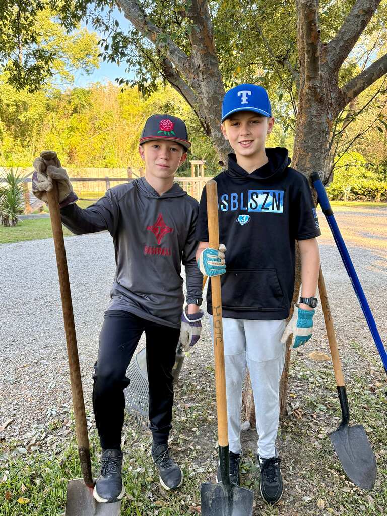 Two boys holding shovels and tools, wearing gloves and caps, standing in a gravel area near a tree.