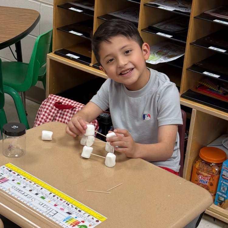 A young boy sits at a desk, smiling, and arranges marshmallows and toothpicks. A ruler is on the desk.