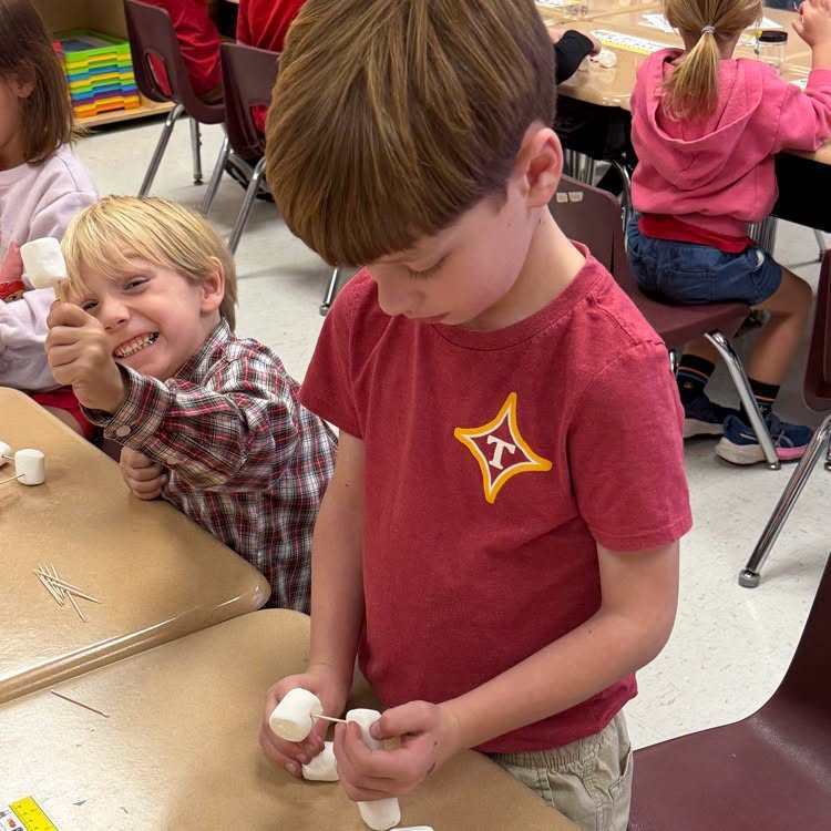 Several students sit at tables, two boys work on small bottles with paint. One boy smiles, another frowns.
