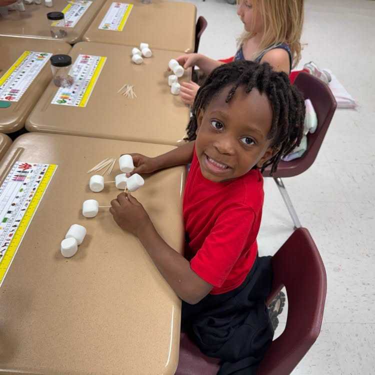 A child in a red shirt arranges small objects on a classroom table while smiling.
