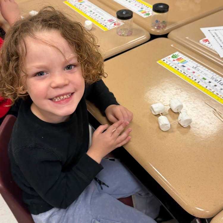 Child sitting at a desk, smiling, with hands on chin; several small white items on desk.