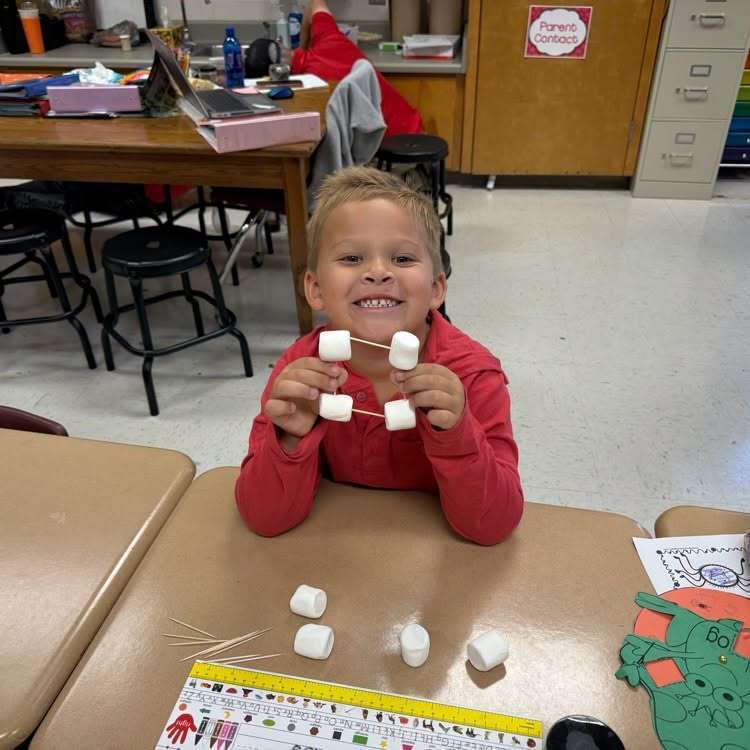 Child seated at a table in a classroom, holding a connected structure made of marshmallows and sticks.