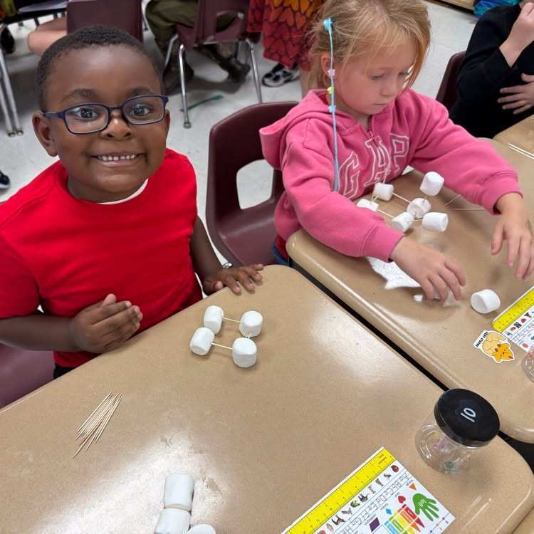 Two children work with marshmallows and toothpicks on desks. A girl smiles; a boy looks at the camera.