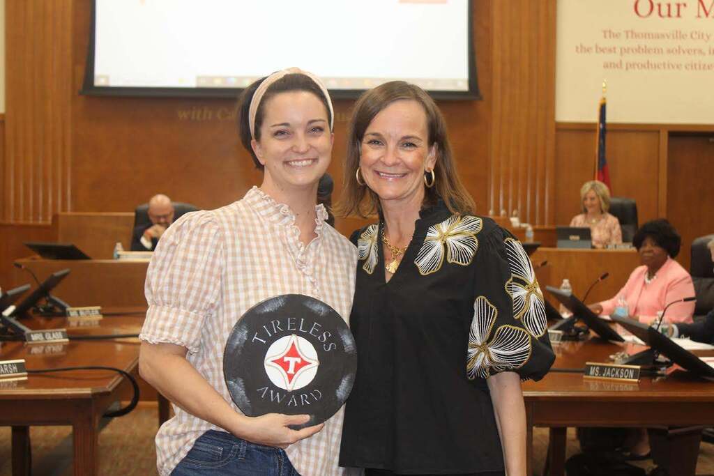 Two women hold a circular award in a large room with a conference table and people sitting.